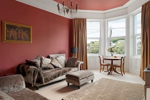 Une pièce avec un mur rouge, un canapé, une table à manger avec des chaises, un lustre et une fenêtre avec des rideaux.