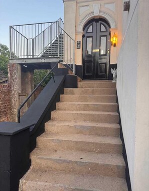 Un conjunto de escalones de piedra que conducen a una puerta negra con una placa de identificación, una pequeña estatua y una lámpara en la pared.