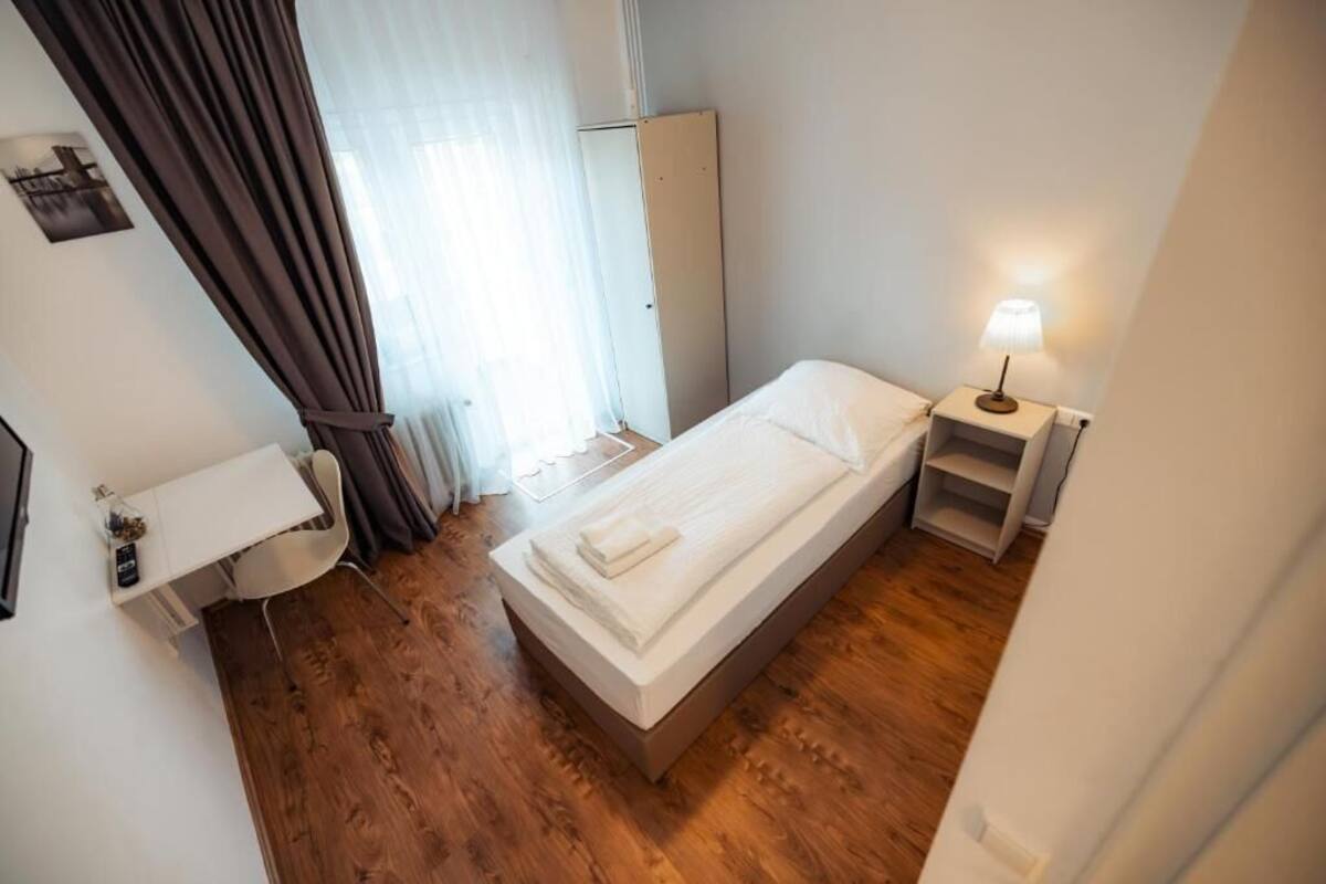 A single bed with a white mattress and pillows, a bedside table with a lamp, a small white desk with a chair, a wardrobe, and a window with curtains.