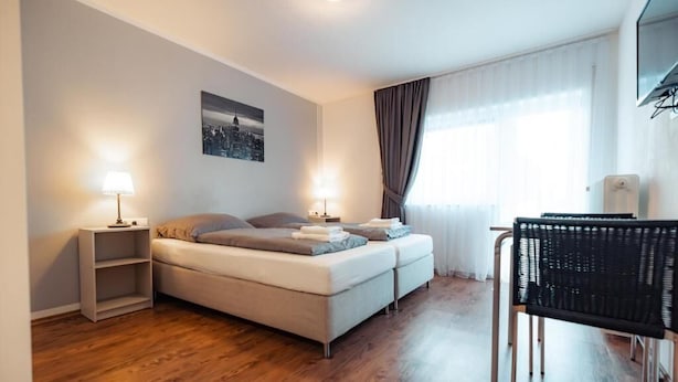 A modern hotel room with a sofa bed, two bedside tables with lamps, a chair, and a cityscape picture on the wall. - Hotel Three Corners (Grenzach-Wyhlen)