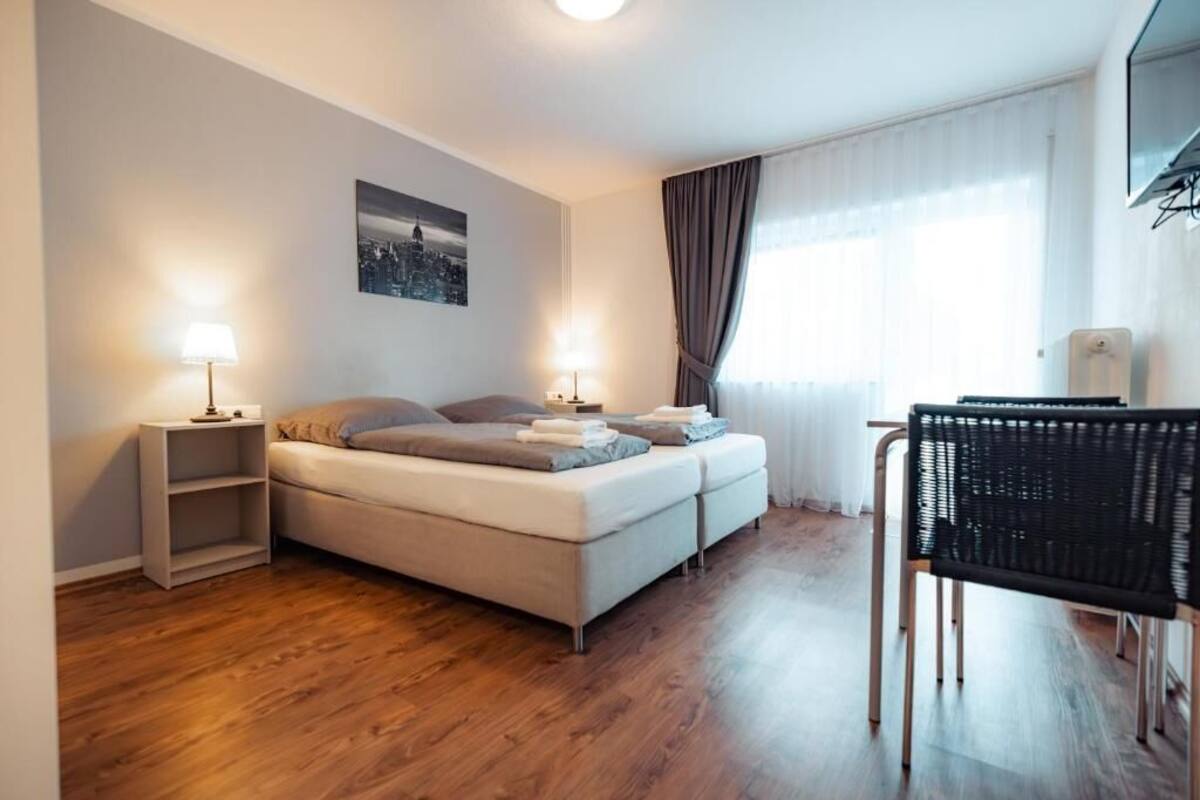 A modern hotel room with a sofa bed, two bedside tables with lamps, a chair, and a cityscape picture on the wall.