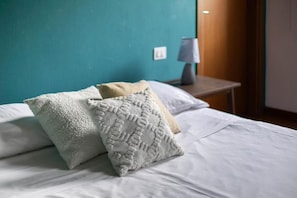 A bed with white bedding and two textured pillows, a bedside table with a lamp, and a teal wall.