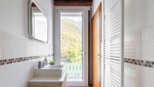 A bathroom with a sink, mirror, and a view of a balcony with a mountain backdrop. - Rabi Rooms (Valbrenta)