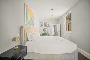 Ein Schlafzimmer mit Bett, Nachttisch, Lampe, Kunstwerk und einem Fenster.