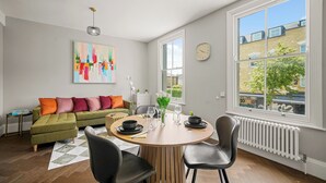 Ett modernt vardagsrum med ett matbord, stolar, en soffa, ett element, en klocka och ett stort fönster med utsikt över byggnader och träd.