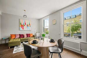Ein modernes Wohnzimmer mit Esstisch, Stühlen, einer Couch, einem Heizkörper, einer Uhr und einem großen Fenster mit Blick auf Gebäude und Bäume.