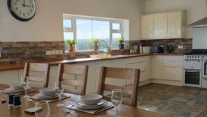 A kitchen with a dining table set for four, a clock, and a view through the window. - Large coastal home in the countryside of Amlwch - Pass the Keys (Amlwch)