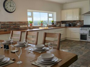 A kitchen with a dining table set for four, a clock, and a view through the window. - Large coastal home in the countryside of Amlwch - Pass the Keys (Amlwch)