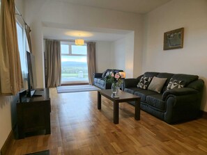 A living room with a sofa, a coffee table, a TV, and a large window with curtains. - Large coastal home in the countryside of Amlwch - Pass the Keys (Amlwch)
