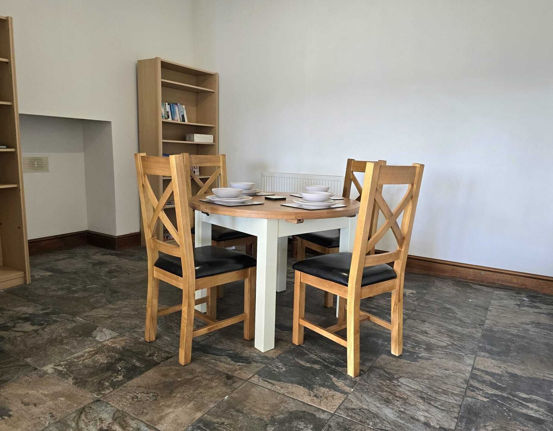 A dining area with a round table, four wooden chairs, and a bookshelf.