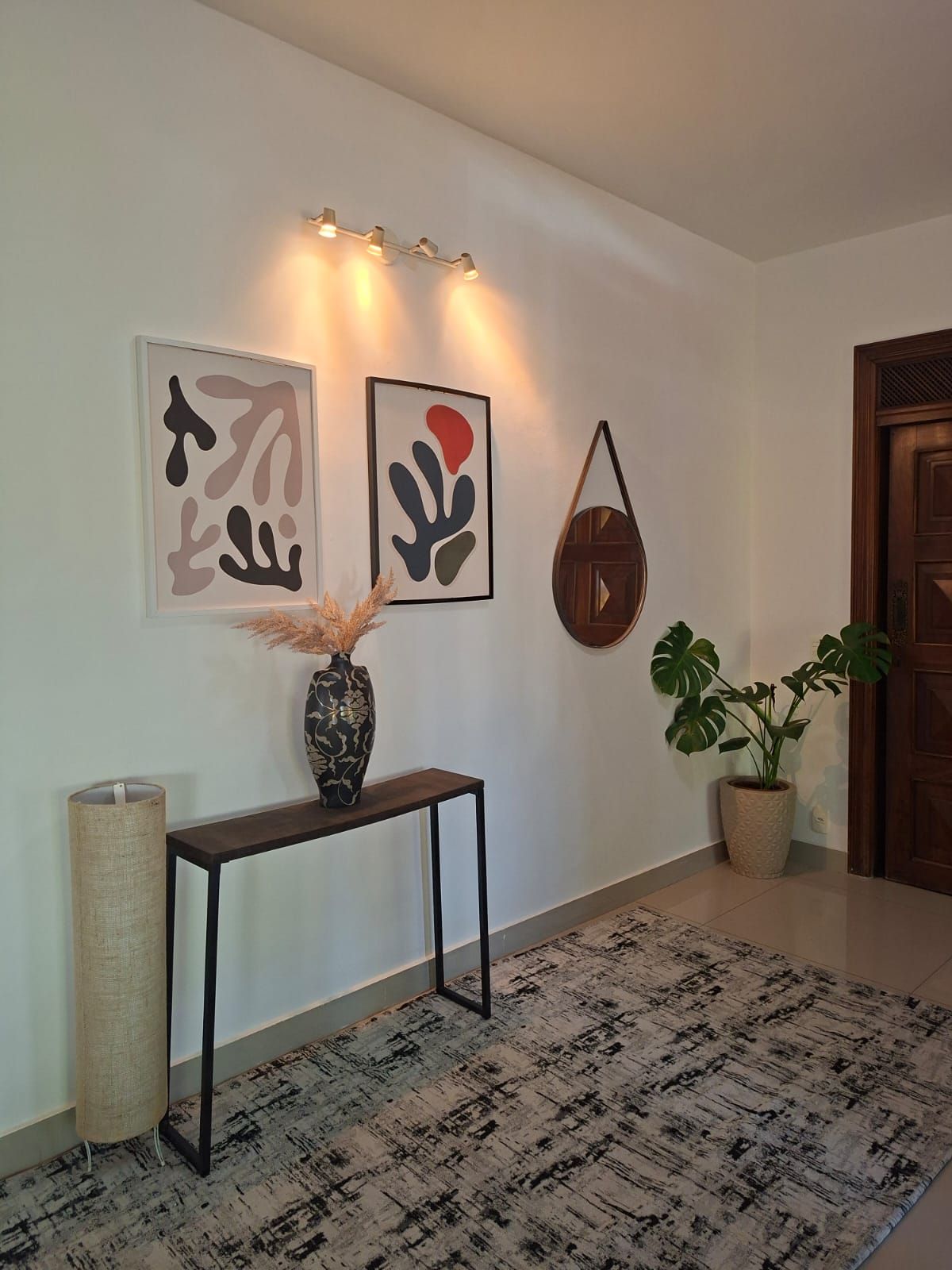 Um corredor com um console de madeira escura, um vaso com plantas secas, arte abstrata na parede, um vaso de planta e um tapete texturizado.