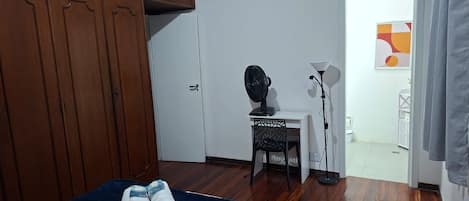 Un dormitori amb un llit, un armari, un escriptori amb una cadira, una làmpada de peu i una porta que dona a una altra estança.