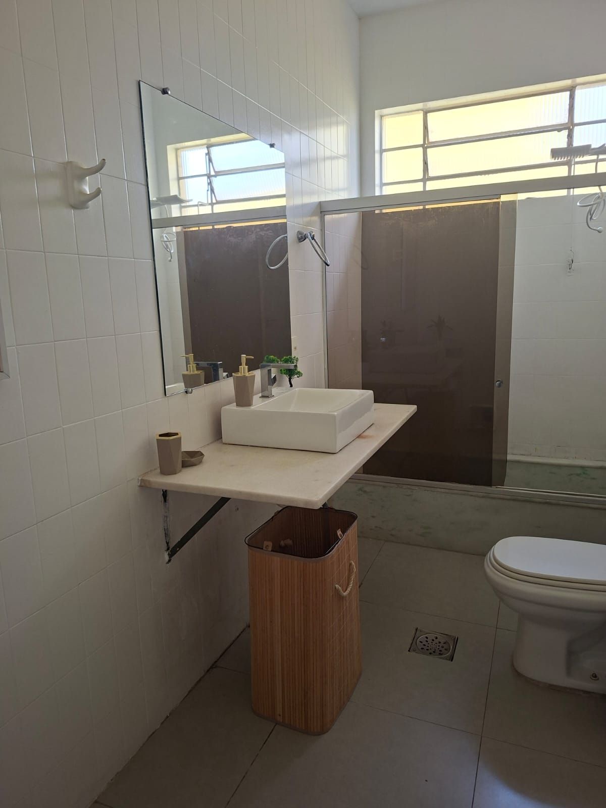 A bathroom with a white sink, mirror, and toilet. There is a wooden trash bin and a window providing natural light.