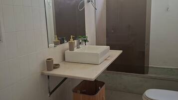 A bathroom with a white sink, mirror, and toilet. There is a wooden trash bin and a window providing natural light.
