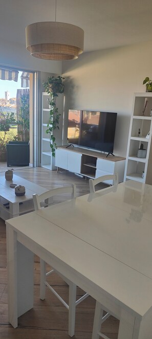 A modern living room with a large flat-screen TV, white furniture, and a large window with a view of buildings and trees. - Fully renovated 3-room apartment, quiet and close to downtown (Vence)
