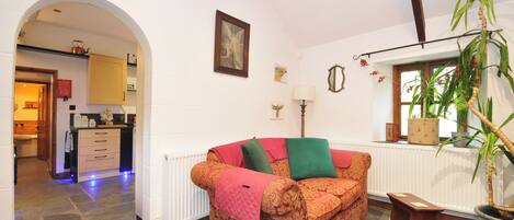 A cozy living room with a patterned sofa, a radiator, a potted plant, and a view of a kitchen through an archway.