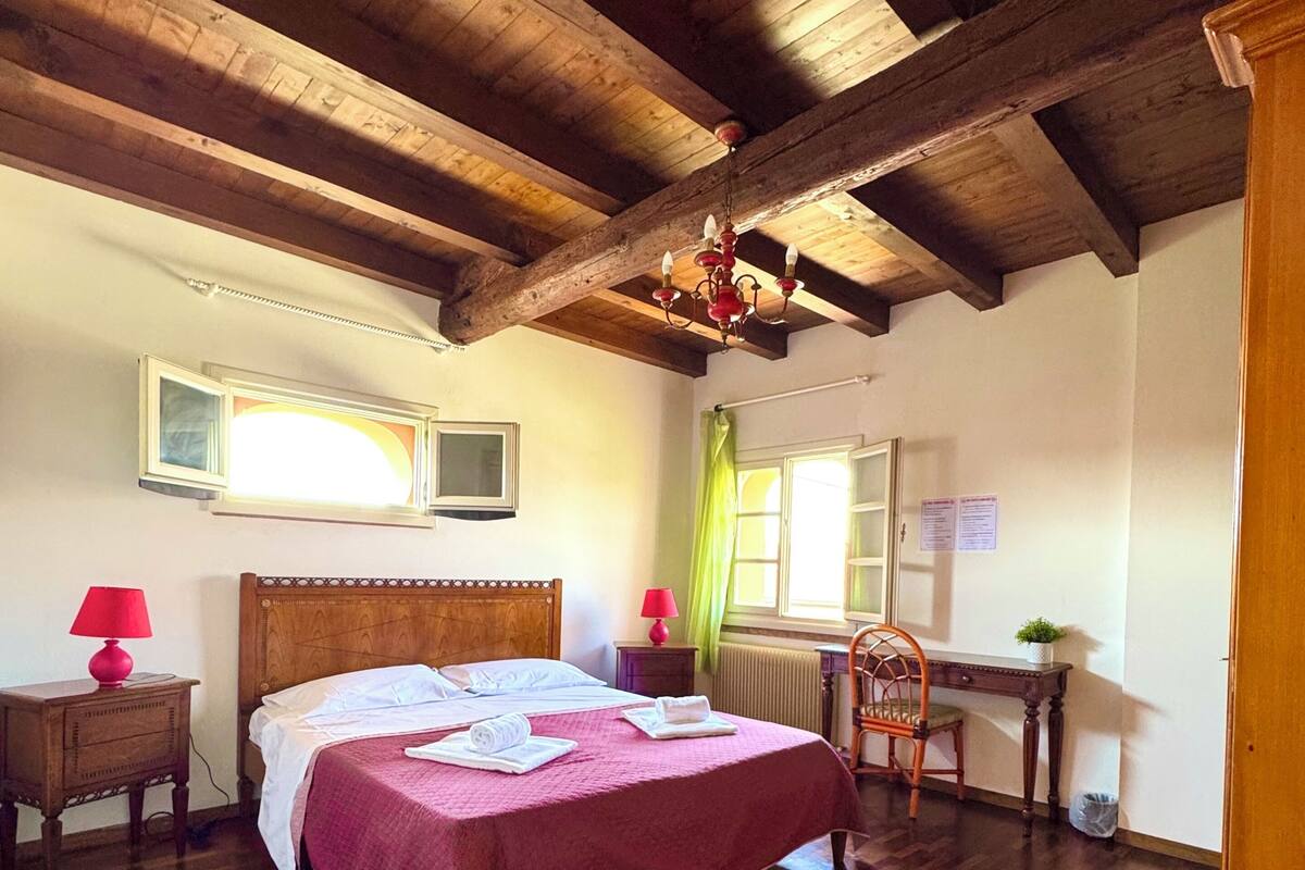 A bedroom with a wooden ceiling, a bed with a red bedspread, a chair, a desk, and a window with curtains.