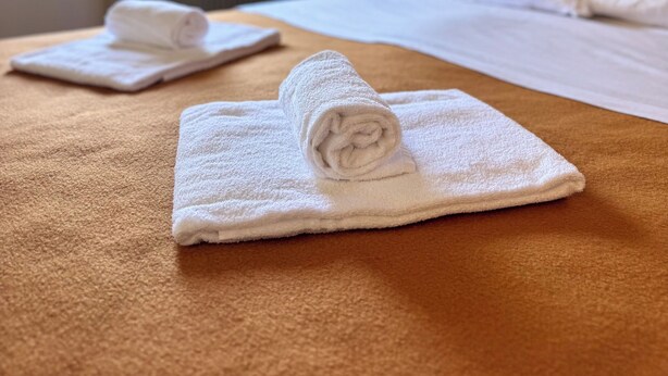 A hotel room with a bed, two rolled towels, a lamp, and a window with curtains. - Biffi Rooms (Bassano del Grappa)
