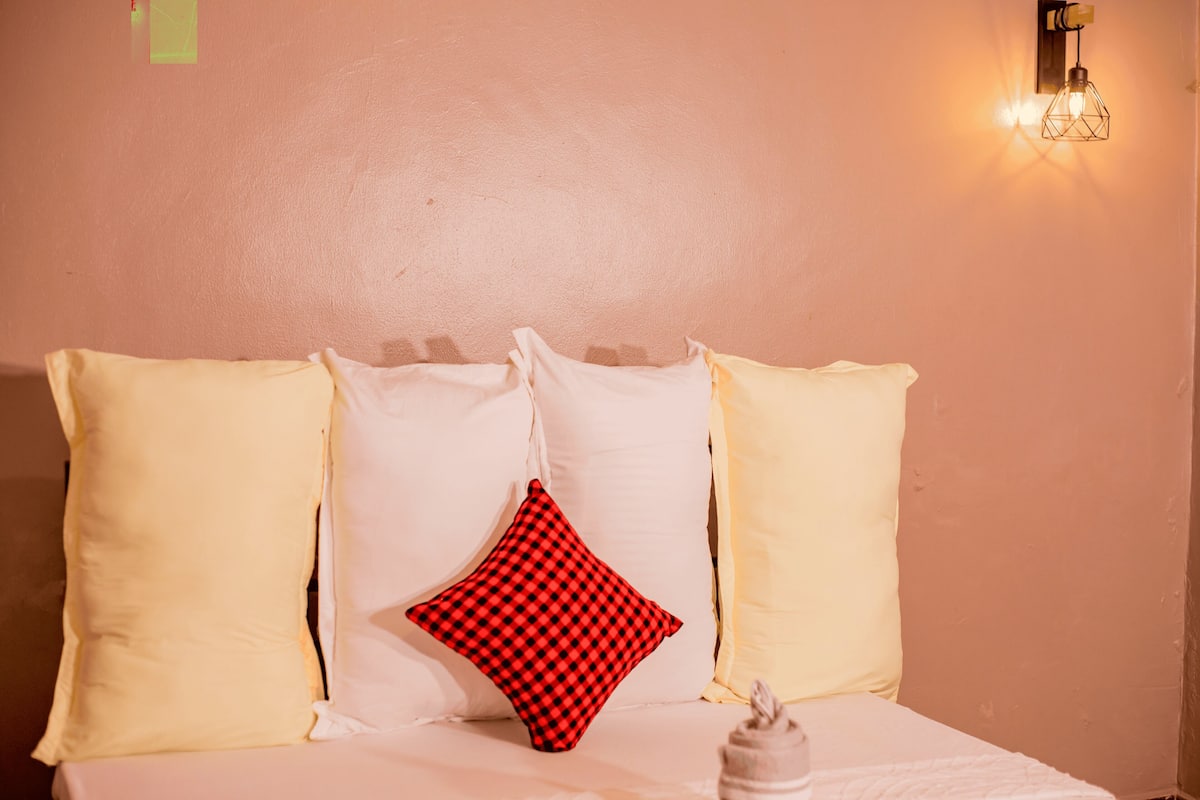 Een bed met witte en gele kussens, een rood-zwart geruit kussen, en een wandlamp.