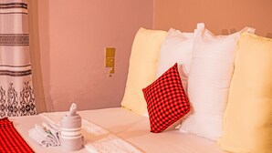 Una cama bien hecha con un cojín de cuadros, una botella blanca y toallas sobre la cama, con una cortina estampada al fondo.