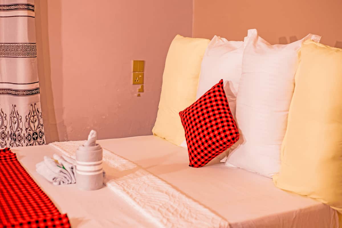 Een net opgemaakt bed met een geruit kussen, een witte fles en handdoeken. Op de achtergrond een bed met een patroon gordijn.