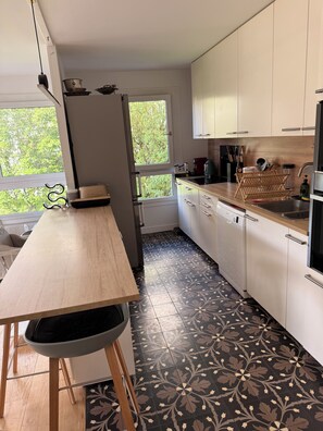 Eine moderne Küche mit einem Holztisch, weißen Schränken, einem gemusterten Boden und einem Fenster mit Blick auf Bäume.