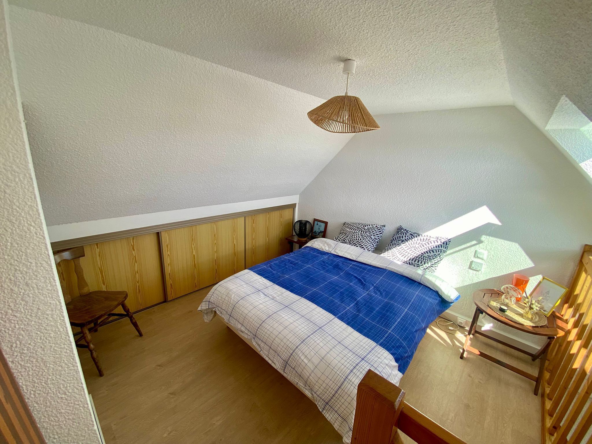 A bedroom with a sloped ceiling, a bed with a blue and white checkered bedspread, a wooden headboard, a bedside table with a lamp, and a window providing natural light.