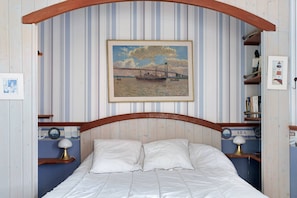 Una cama con cabecera de madera, un cuadro enmarco de un puente y una pequeña estantería con una lámpara.