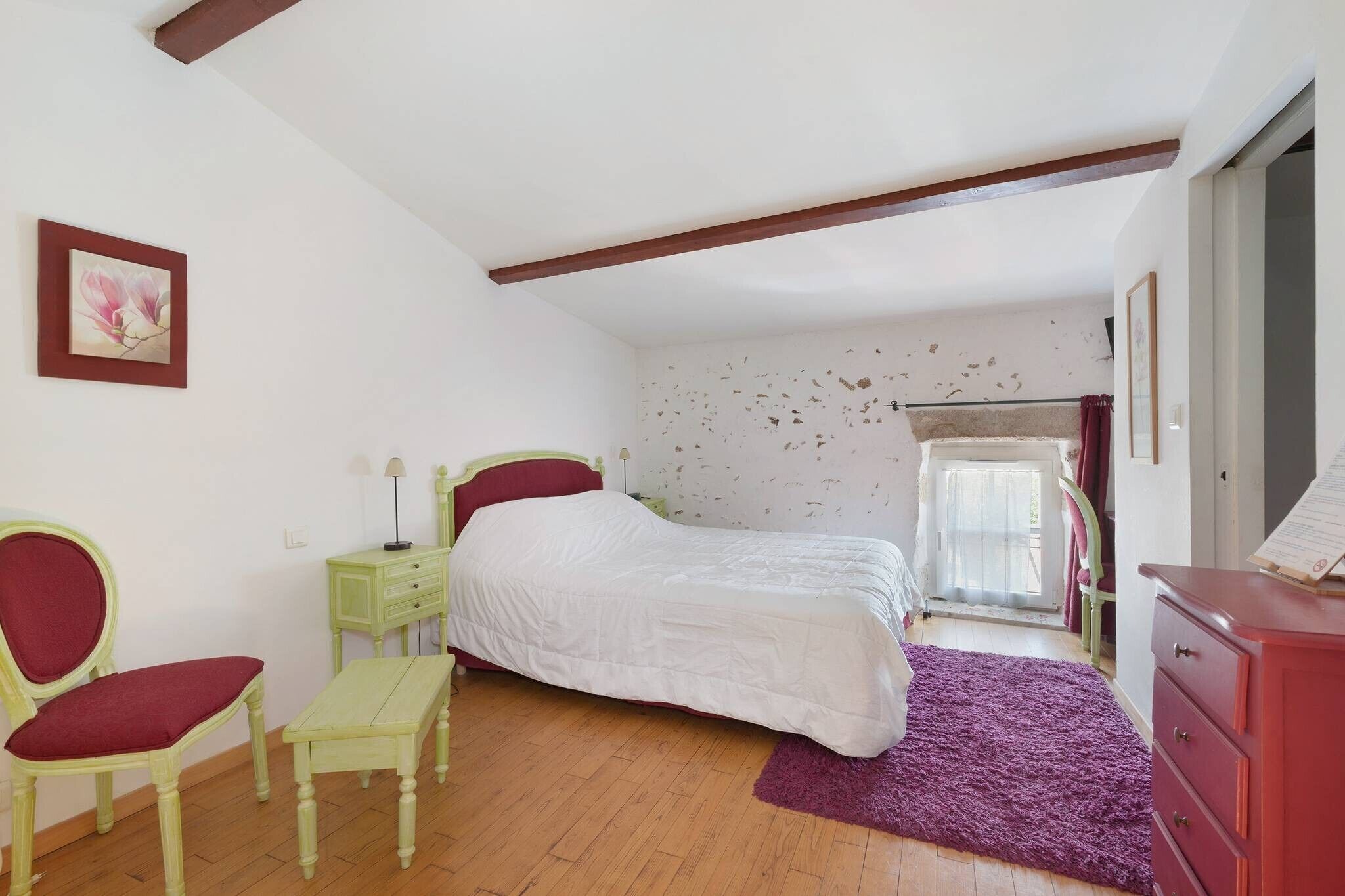 Un dormitorio con cama, una silla, una mesita, un tocador y una ventana con cortinas.
