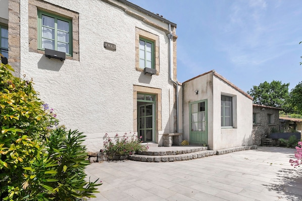 Un bâtiment à deux étages avec une façade en pierre, une petite construction attenante dotée d’une porte verte et une cour pavée.