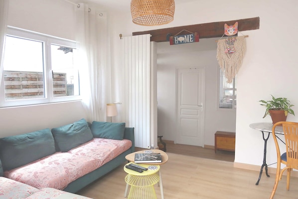 Ein Zimmer mit einer Couch, einem kleinen Tisch mit Fernbedienung, einem Sessel, einer hängenden Makramee-Dekoration und einem Schild mit der Aufschrift 'HOME'.