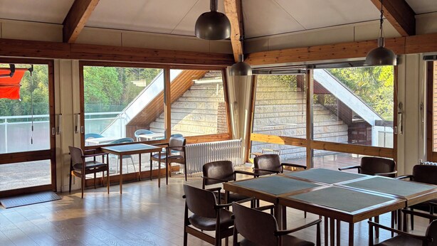 Ruime kamer met hoge plafonds, houten balken, grote ramen en een tafel met stoelen.