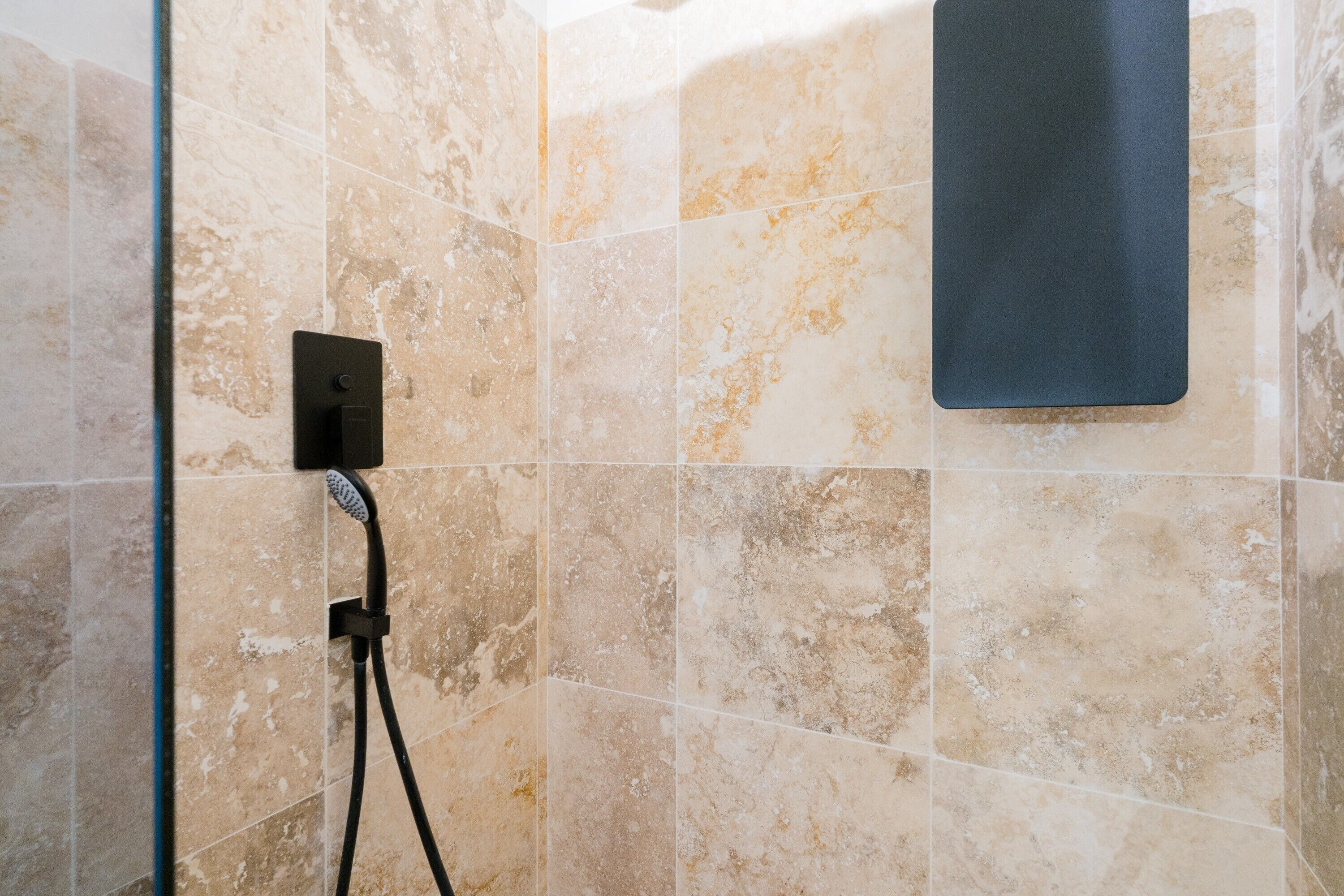 A bathroom with beige marble tiles, a shower head, and a black control panel.