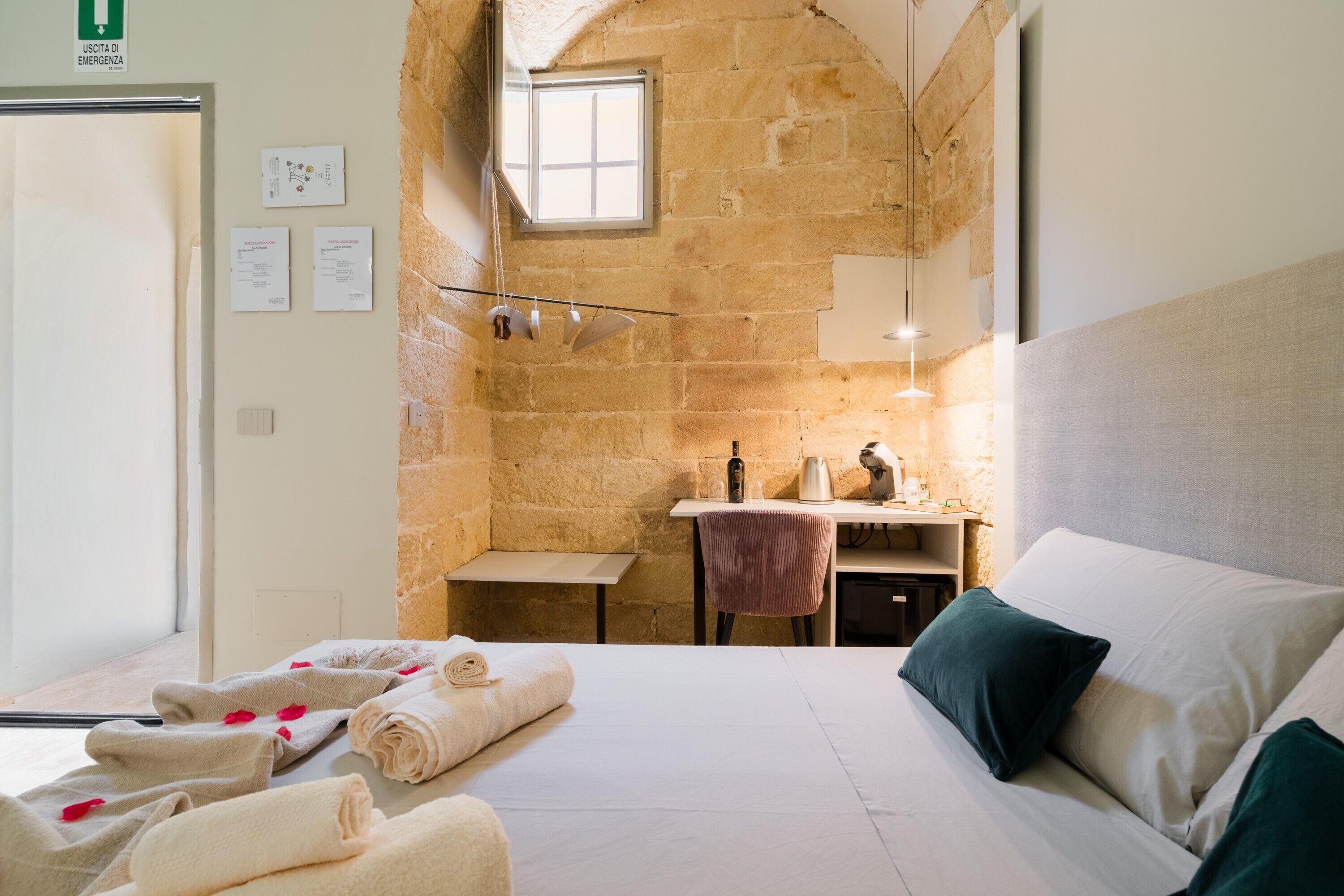 Quarto de hotel com uma cama, uma escrivaninha, uma cadeira, uma mesinha, uma janela e um sinal de saída de emergência fixado na parede.