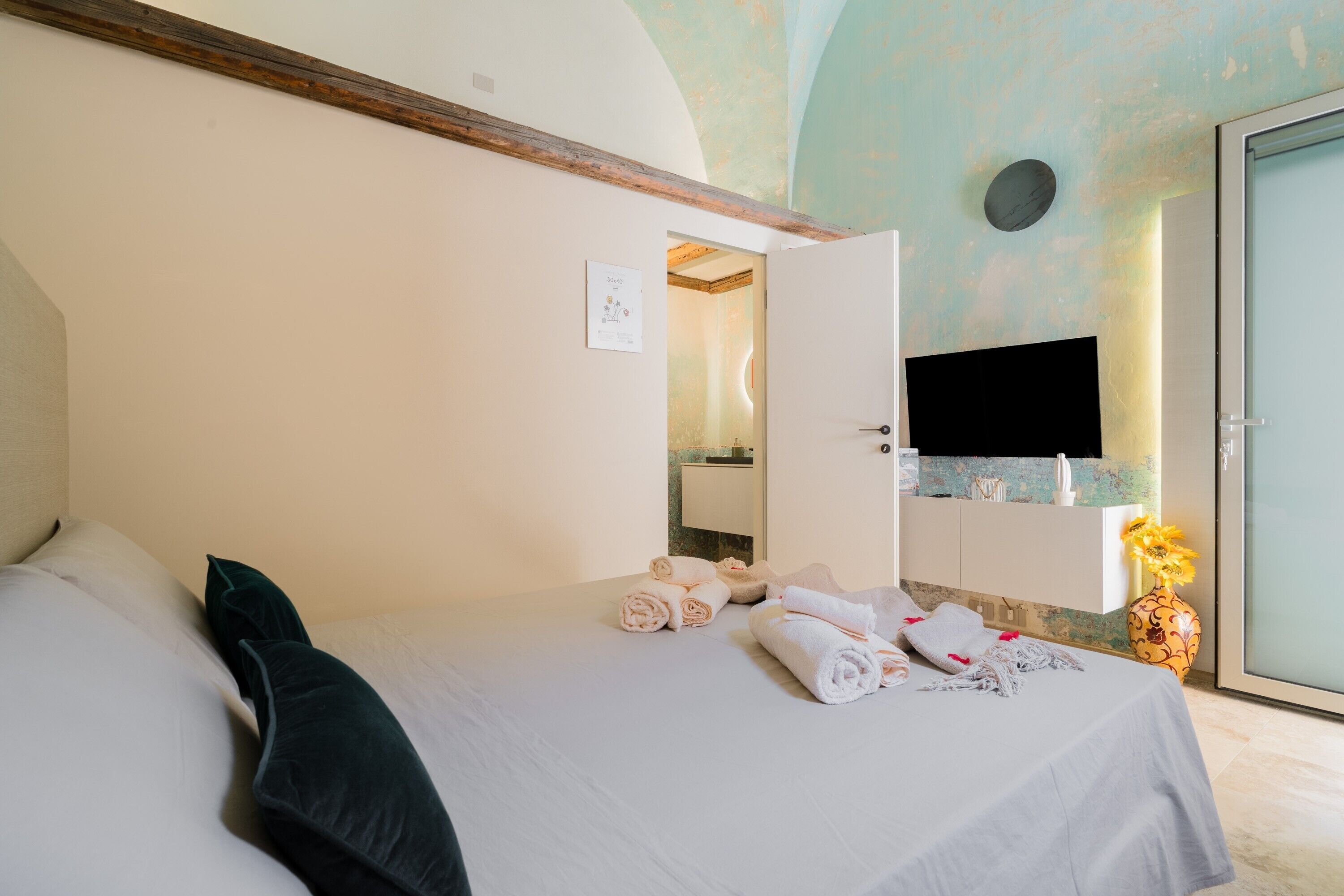 Una camera d'albergo con un letto, uno specchio nel bagno, un lavandino e un vaso con fiori.