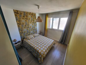 A bedroom with a bed, a hanging light fixture, a window with curtains, and a wall with a tropical pattern. - Bright apartment in Toulon (Toulon)