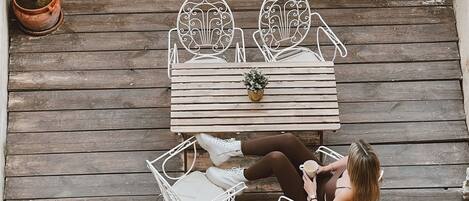 Une terrasse en bois avec une table et des chaises, une plante en pot et une personne assise.