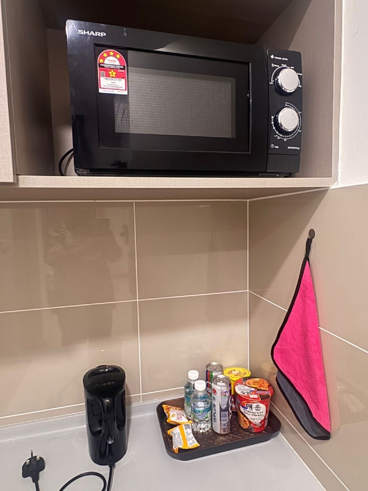 Eine Mikrowelle auf einem Regal, ein Tablett mit Snacks und Getränken sowie ein pinkes Handtuch an der Wand.
