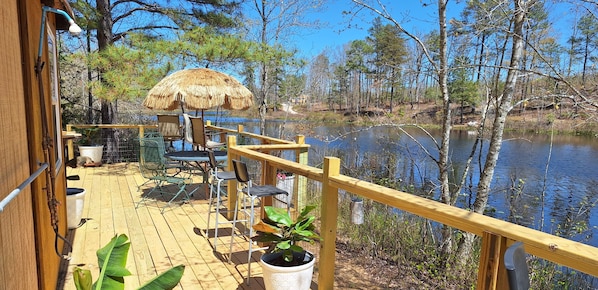 A wooden deck with outdoor furniture, a thatched umbrella, and a view of a lake surrounded by trees.