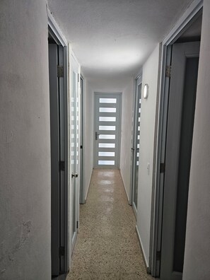 A narrow hallway with doors on both sides, a tiled floor, and a door at the end with horizontal frosted glass panels. - Cozy 3 BR Luquillo bungalow. Close proximity to beach, kiosks, and rainforest. (Luquillo)