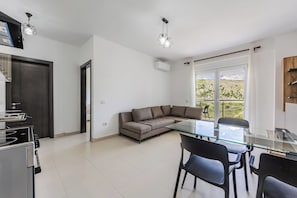 A modern kitchen with a built-in stove, a dining area with a glass table and chairs, a sofa, and a large window with a view of greenery.