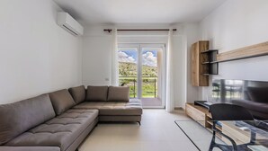 Ein modernes Wohnzimmer mit einer Eckcouch, einem Flachbildfernseher und einem Balkon mit Blick auf Grünflächen.