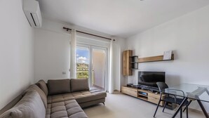 Ein modernes Wohnzimmer mit einer Eckcouch, einem Glastisch, einem Fernseher und einer Schiebetür, die auf einen Balkon führt.