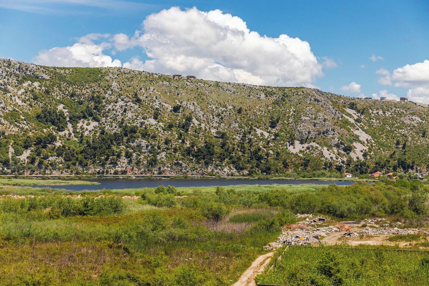 Paisaje montañoso con un lago, un sendero de tierra y edificios dispersos.
