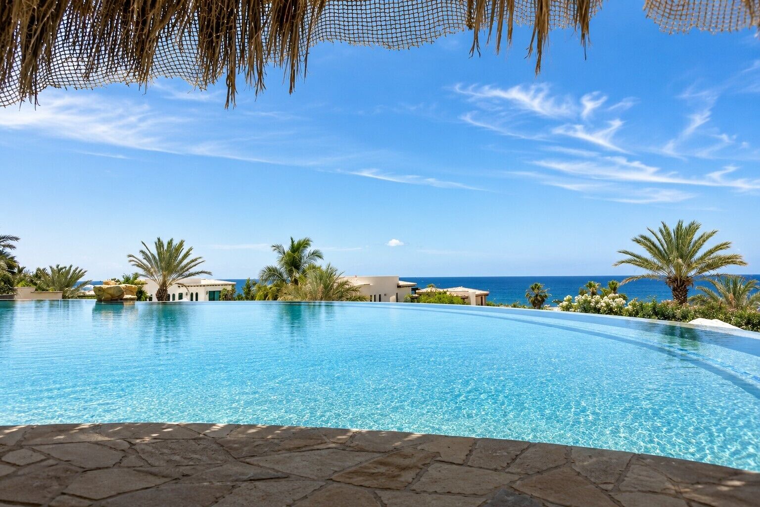 A clear blue pool with a view of the ocean, surrounded by palm trees and buildings.