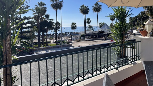 Un balcón con vista al mar, palmeras y una calle con un café.