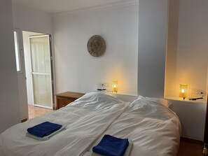 Ein Schlafzimmer mit einem Bett, zwei Nachttischlampen, einem kleinen Holfnachtisch und einer Tür zu einem anderen Raum.