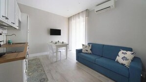 A modern living space with a kitchenette, a dining area, a blue sofa, and a mounted TV. - Hostdomus - Stella Maris (Alassio)