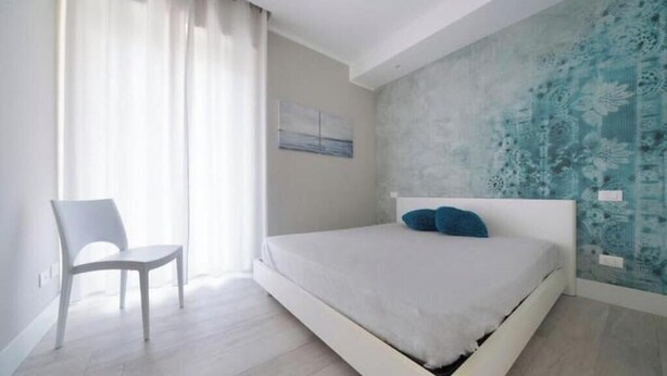 A bedroom with a bed, a chair, a window with curtains, and a wall with a floral pattern. - Hostdomus - Stella Maris (Alassio)