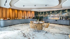 A modern dining area with a round table, wicker chairs, a marble floor, and a kitchen counter with various food items. - Green Bay Thassos (Thasos)